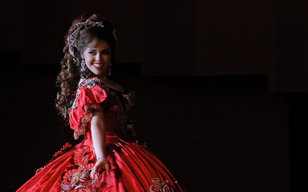 Film still of young Latina smiling with large curly hairstyle and wearing a red colonial style dress.