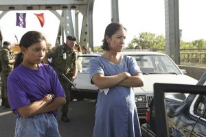A woman and young girl both cross their arms looking at a car. A military person is in the background.