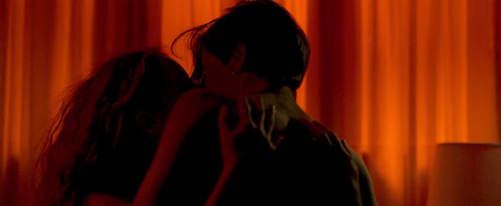 Two people embrace in a dim lit room with red curtains.