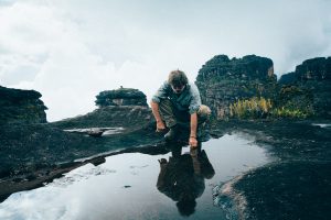 A man bends over to touch a puddle of water among rock formations. His reflection is seen on the glossy water.