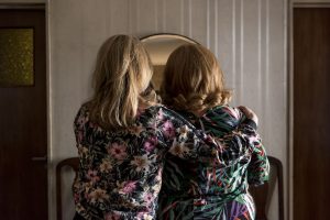 Two women with blonde hair face away towards a mirror. Both are wearing jackets with pink flowers.