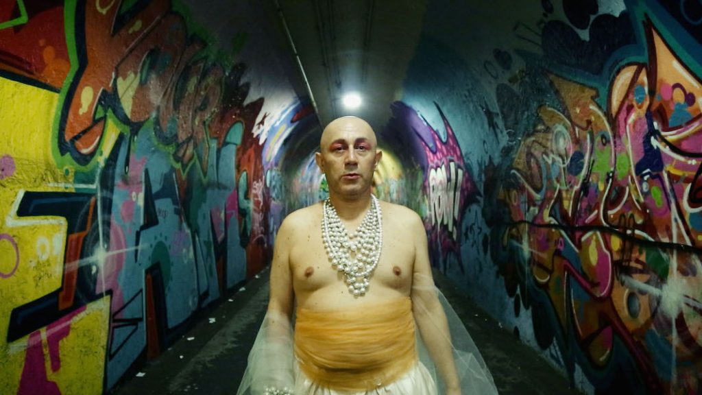 A transgender individual wearing necklaces and a yellow skirt standing in a tunnel with graffiti.